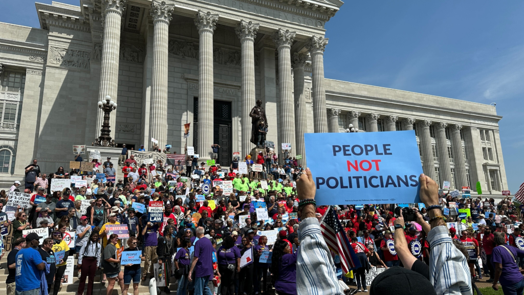 Thousands of people stand in front of the Missouri capitol building holding signs demanding an end to democracy. In the foreground hands hold a sign saying People Not Politicians.